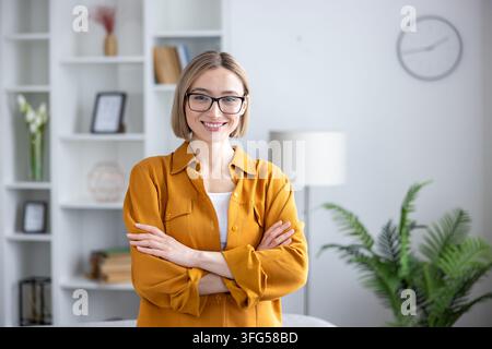 Jeune femme confiante debout les bras croisés. Portant des lunettes et une chemise jaune. Souriant à l'appareil dans un bureau ou espace de travail moderne. Représentant le professionnalisme. Le succès. Et le leadership Banque D'Images
