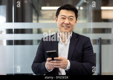 Homme d'affaires asiatique dans un bureau moderne souriant avec confiance à la caméra tout en tenant un smartphone, exprimant le succès professionnel, le réseautage, la technologie et la vie d'entreprise contemporaine Banque D'Images