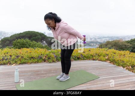 Femme afro-américaine âgée se penchant sur un tapis vert sur le pont, vêtue d'une veste rose tenant un haltère violet Banque D'Images
