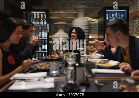 Un groupe d'amis divers savoure de la nourriture et des boissons tout en ayant des conversations animées dans un restaurant en soirée. Banque D'Images
