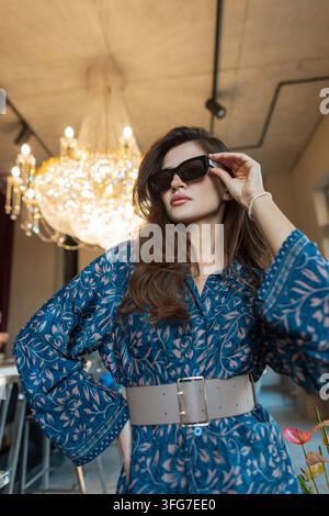 jeune femme confiante pose dans une robe bleue à motifs et une large ceinture, ajustant ses lunettes de soleil. Prise de vue en contre-plongée dans un salon moderne avec un lustre en cristal allumé Banque D'Images