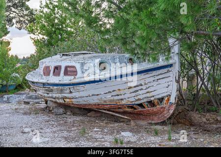 Un bateau en bois abandonné avec de la peinture écaillée et un coque rouillée repose sur une rive rocheuse parmi des pins, évoquant la décrépitude, la nostalgie et l'abandon côtier Banque D'Images