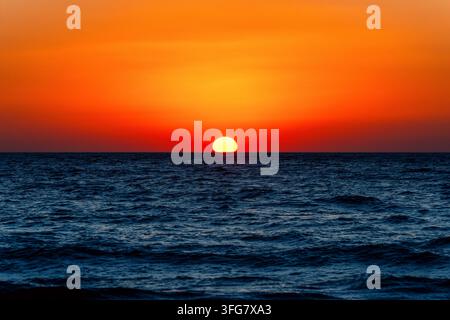 Le soleil éclatant touche l'horizon de la mer au coucher du soleil au large de Crète, en Grèce, sous un ciel orange et rouge intense au-dessus d'ondes bleu foncé et d'un paysage marin calme du soir Banque D'Images