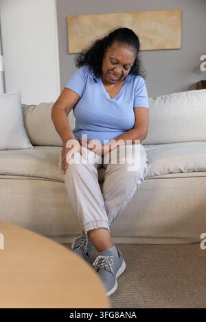 Sénior afro-américaine assise sur un canapé beige massant son genou droit avec des baskets grises visibles. Confort, mobilité, soins à domicile, cosy, bien-être, agi Banque D'Images