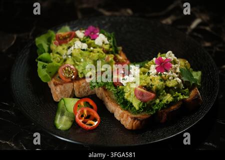 Deux tranches de pain rustique garnies d'avocat, de légumes verts, de tomates cerises, de fromage effrité et de fleurs comestibles sur une assiette noire foncée. Banque D'Images