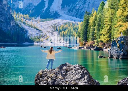Vue arrière d'une femme debout les bras ouverts sur un point de vue rocheux au-dessus du turquoise lac de Braies, embrassant le paysage alpin. Idéal pour les campagnes sur la liberté, le bien-être, les voyages en solo, le tourisme et le style de vie en plein air Banque D'Images