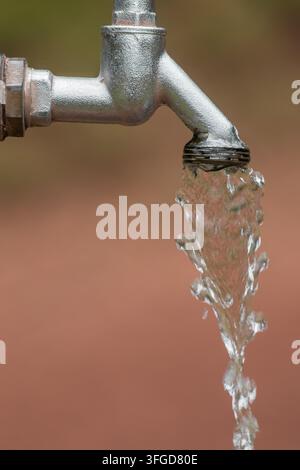 Un robinet laisse couler un jet d'eau dans un espace extérieur. L'eau cascade du bec en métal sur le sol sec. La lumière du soleil se reflète sur le fl Banque D'Images