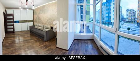 Intérieur d'appartement moderne avec canapé gris, armoire coulissante et fenêtres panoramiques donnant sur des bâtiments résidentiels. Espace de vie élégant avec sol en bois et concept de design contemporain Banque D'Images