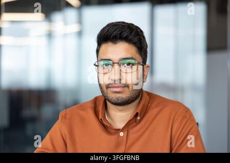 Portrait rapproché d'un homme en chemise couleur rouille avec des cheveux foncés, des lunettes et une barbe courte, assis dans un intérieur de bureau moderne avec un fond flou doux, expression calme, espace de copie Banque D'Images
