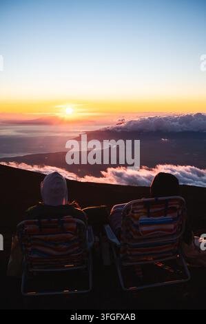 Couple se relaxant dans des chaises longues et regardant le coucher de soleil au-dessus des nuages depuis le sommet du volcan Haleakala, Maui, Hawaï, États-Unis Banque D'Images