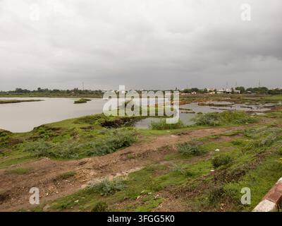 Un chemin rural serein longe une rivière calme sous un ciel nuageux au Gujarat, en Inde. Le cadre tranquille est idéal pour dépeindre des thèmes de la nature Banque D'Images