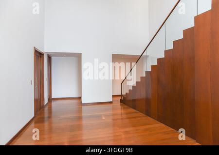 Couloir moderne avec escalier en bois : Un couloir contemporain mettant en vedette un escalier en bois frappant avec une rambarde en verre, menant aux étages supérieurs, illuminé Banque D'Images