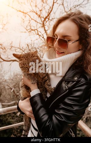 Femme en veste en cuir de chat Jeune femme élégante tenant un chat tigré dans un parc automnal tout en portant des lunettes de soleil tendance Banque D'Images