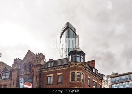 Vue de la façade moderne en verre du Radisson Blu Hotel s'élevant derrière des bâtiments historiques en briques rouges sous un ciel nuageux à Birmingham, Angleterre, Royaume-Uni Banque D'Images