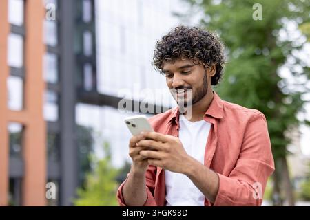 Jeune étudiant universitaire souriant marchant sur le campus en utilisant son smartphone pendant la pause. Banque D'Images
