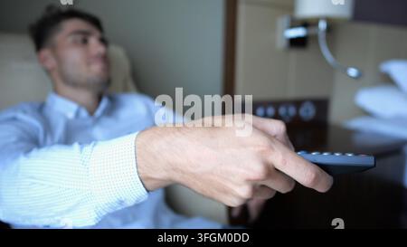Focus sur la main masculine du jeune homme d'affaires dans les canaux de commutation de fauteuil sur la télécommande de télévision à la chambre d'hôtel moderne. Détente cosy lors d'un voyage d'affaires Banque D'Images