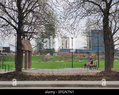 Une vue pittoresque d'un parc urbain encadré par deux grands arbres déciduels au premier plan. Au plan moyen, deux personnes sont assises sur un banc en bois du parc Banque D'Images