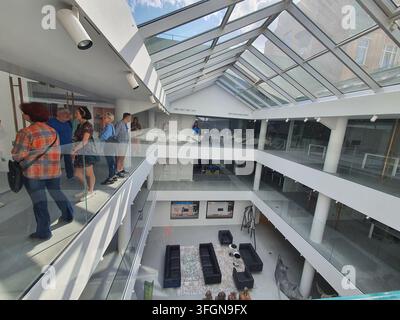 Un intérieur d'atrium moderne à plusieurs niveaux avec un plafond en verre permettant à la lumière naturelle du jour d'inonder l'espace. Plusieurs visiteurs se tiennent sur un niveau supérieur Banque D'Images