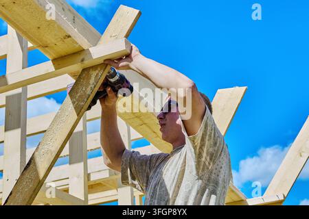 Un ouvrier du bâtiment utilise une perceuse sans fil pour fixer un support en bois lors de l'assemblage d'un cadre en bois à l'extérieur. Banque D'Images