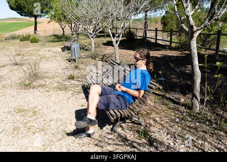 Une jeune personne se détend sur un banc en bois dans un parc sous le soleil. Des arbres et de l'herbe sont à proximité. Banque D'Images