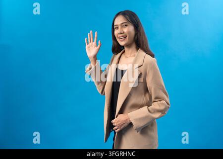 Femme d'affaires indonésienne souriante portant un blazer beige et faisant un signe de la main contre un fond bleu vif du studio avec espace de copie. Banque D'Images