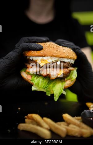 Une personne portant des gants noirs tient un burger empilé avec des couches de viande, de fromage fondu, de laitue et d'autres ingrédients. Des frites croustillantes sont posées sur la table Banque D'Images