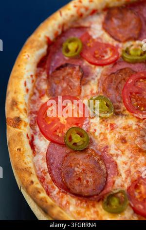 Une pizza ronde repose sur une planche en bois avec des tranches de pepperoni, de tomate fraîche et de jalapeno. La pizza est placée dans un cadre décontracté adapté pour d Banque D'Images