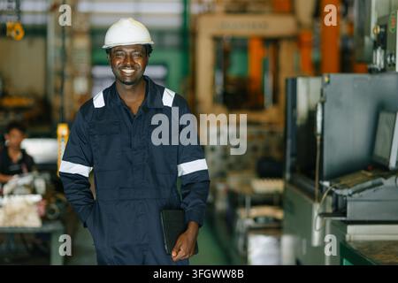 Portrait d'un jeune travailleur noir africain heureux portant un uniforme, une salopette et un casque, souriant à l'appareil à l'intérieur d'une usine de fabrication moderne Banque D'Images