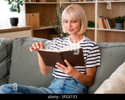 Femme blonde détendue allongée sur un canapé et lisant un livre. Banque D'Images