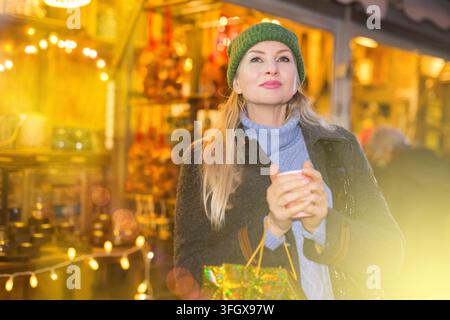 Portrait d'une femme blonde heureuse et attirante visitant un marché de Noël traditionnel en ville, marchant parmi les étals de commerce avec un gobelet en papier de café chaud Banque D'Images