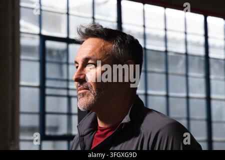 Homme d'âge moyen debout dans un loft industriel près de fenêtres métalliques à multiples panneaux, portant un t-shirt rouge à col rond Banque D'Images