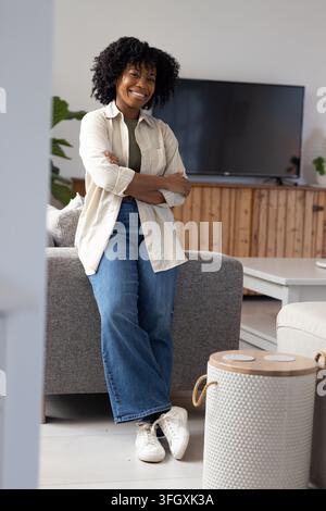 Femme africaine appuyée sur un canapé gris dans le salon, portant un jean et des baskets blanches Banque D'Images