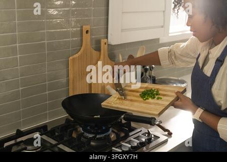 Femme africaine portant un tablier ajoutant des oignons, de l'ail dans un wok dans la cuisine sur une flamme bleue, espace libre Banque D'Images