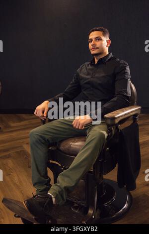 Homme adulte assis dans un fauteuil de coiffeur vintage dans un coin du studio, portant une chemise noire et une veste sombre Banque D'Images