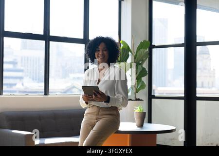 Femme afro-américaine appuyée à une table dans un bureau tenant une tablette, portant un chemisier blanc et un pantalon beige Banque D'Images