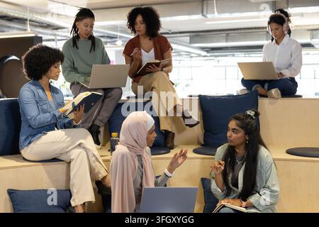 Équipe diversée assise sur des gradins à coussins bleus dans un bureau ouvert, utilisant des ordinateurs portables pour prendre des notes Banque D'Images