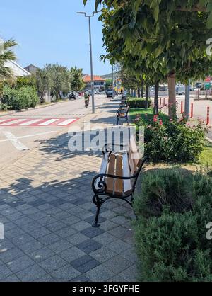 Pirovac, Croatie, 16 août 2026 Rue de quartier calme baignée de soleil avec bancs en bois, cycliste, arbres taillés, fleurs roses, pavage de trottoir, carrefour Banque D'Images