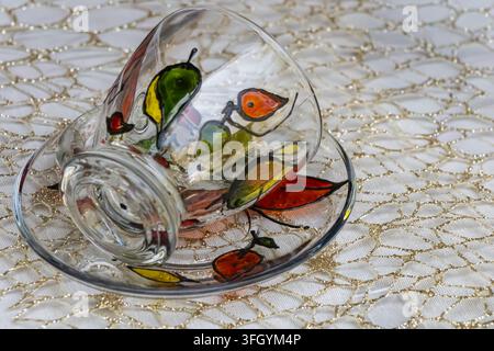Verre décoratif et soucoupe avec des motifs de fruits peints à la main et colorés, disposés sur une nappe blanche et dorée à motifs dans un élégant décor de table Banque D'Images