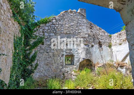 Ruines d'une maison en pierre abandonnée avec des murs couverts de lierre et une cour envahie par la végétation, baignées de soleil sous un ciel bleu clair. Maçonnerie érodée, une fenêtre brisée et Banque D'Images