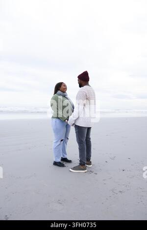 Couple africain tenant la main, debout sur une plage de sable mouillée, portant un bonnet et montrant des empreintes de pas Banque D'Images