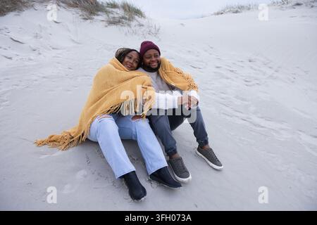 Couple assis enveloppé dans une couverture jaune moutarde sur des dunes de sable, portant un bonnet marron Banque D'Images
