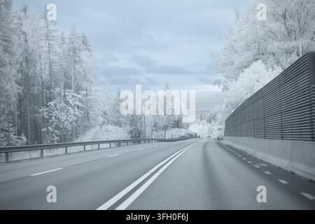 Une route s'étend à travers des arbres enneigés dans un environnement froid. La scène montre un chemin lisse avec un garde-corps à côté. Le ciel est nuageux, typique pour w Banque D'Images