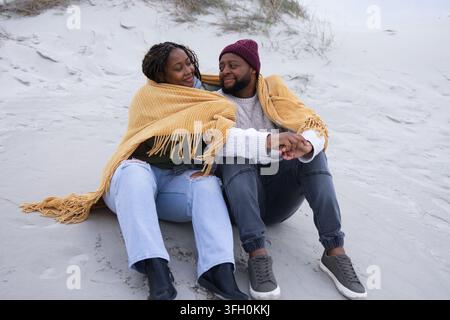 Couple africain assis sur une dune côtière enveloppé dans une couverture moutarde, se tenant la main Banque D'Images