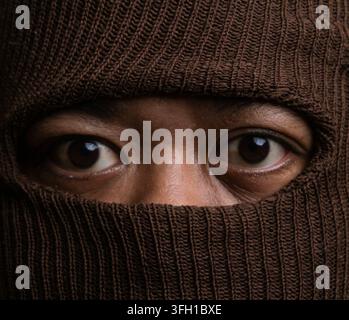 Portrait rapproché captivant montrant les yeux sombres perçants d'un homme indonésien, encadrés de près par un passe-montagne tricoté marron. Matériau de laine épais Banque D'Images
