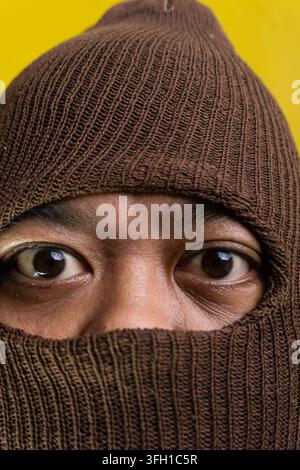 Portrait macro offrant un gros plan extrême des yeux sombres et intenses appartenant à un homme asiatique, complètement englouti par une cagoule tricotée brune. Épais r Banque D'Images