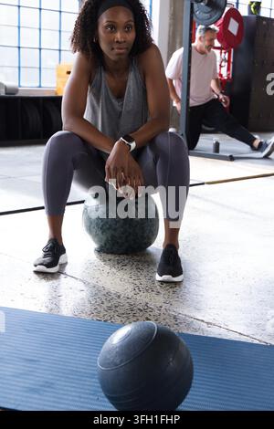 Diverses personnes de gym en vêtements de sport assises sur un ballon de médecine et en position de fente près du rack à squats dans la salle de sport Banque D'Images