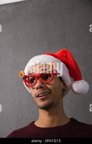 Homme afro-américain portant un chapeau du Père Noël, des lunettes pailletées et un haut bordeaux, regardant vers le haut en studio Banque D'Images