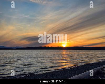 Un coucher de soleil doré brille au-dessus d'une ligne d'horizon lointaine alors que des nuages striés dramatiques remplissent le ciel et que la lumière chaude se reflète sur l'eau ondulante vers une rive rocheuse Banque D'Images