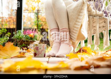 Une femme d'âge moyen se détend sur une terrasse d'automne, les pieds dans des chaussettes en laine chaudes à côté d'une tasse de café et de feuilles colorées tombées, un moment de bien-être cosy. Banque D'Images