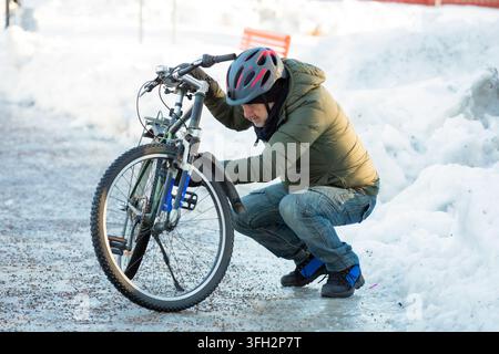 Un homme âgé portant un casque s'accroupit pour inspecter sa roue de vélo sur une route glacée à Helsinki, en Finlande, lors d'une réparation de vélo en hiver. Banque D'Images